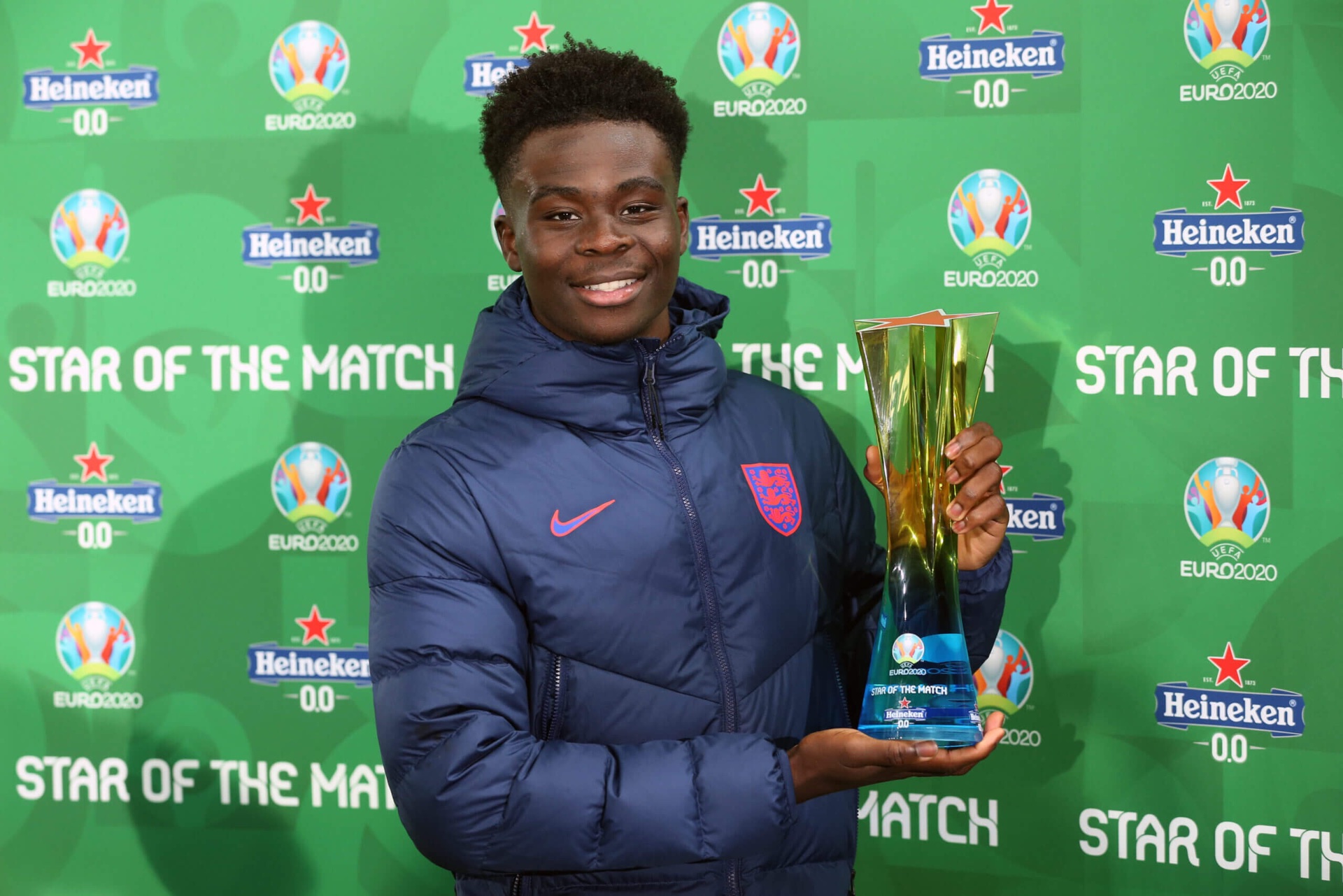 Bukayo Saka smiling in an England jacket while holding a UEFA EURO 2020 Star of the Match trophy.