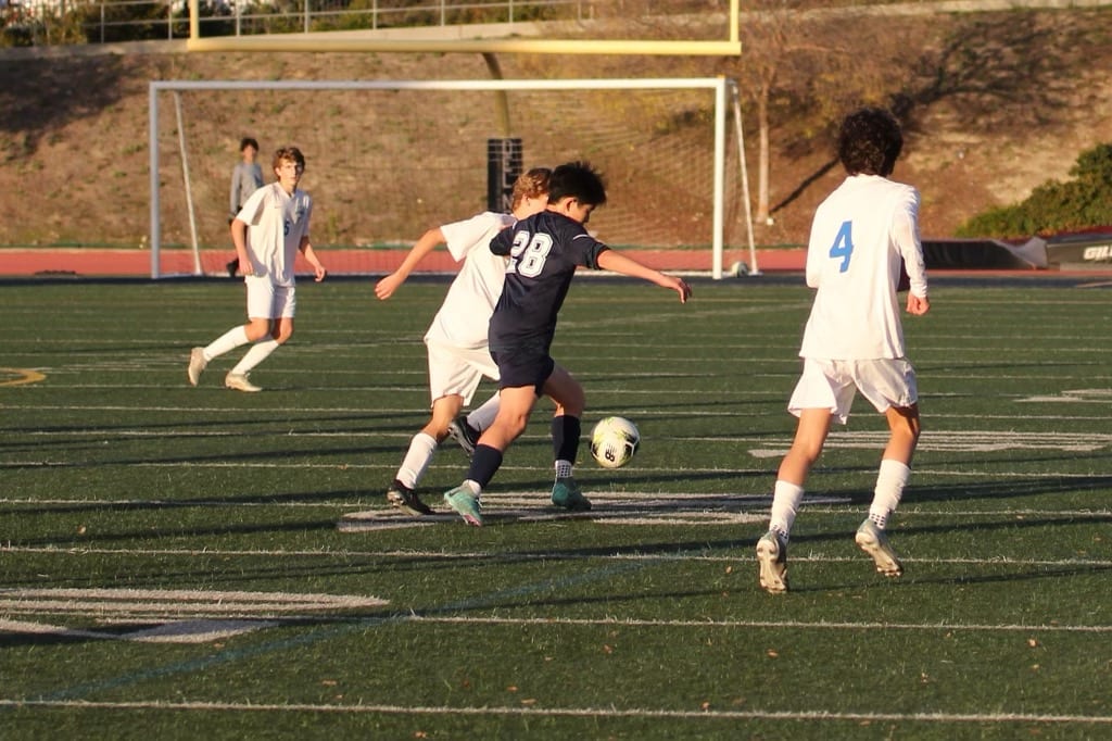 High school soccer player in a dark jersey dribbling between defenders during a game