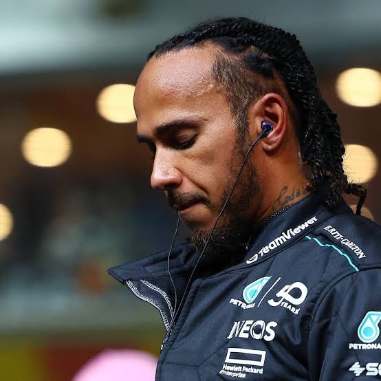 Sir Lewis Hamilton looking downward in reflective focus while wearing his Formula 1 team jacket and earpiece