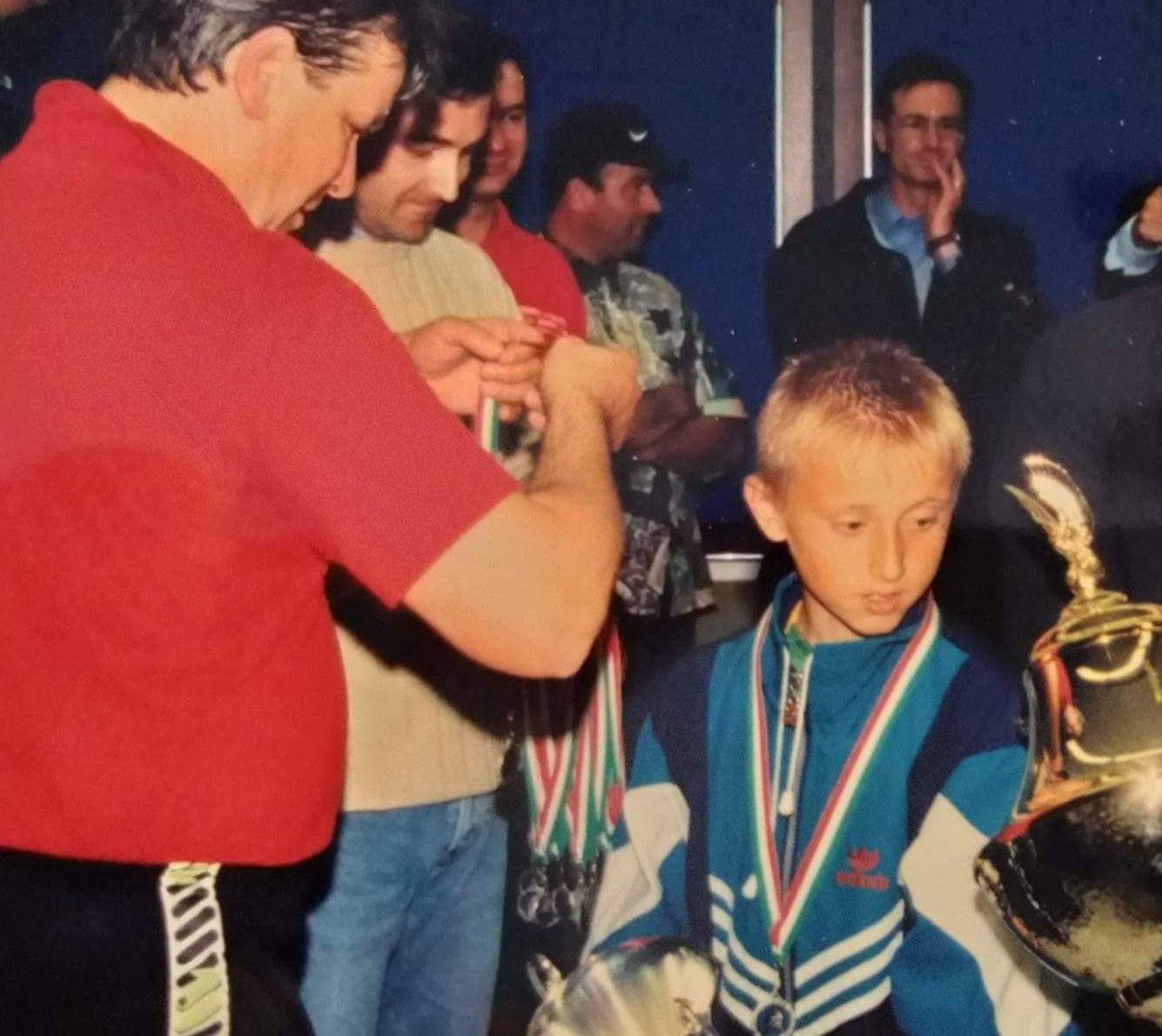 A young Luka Modrić wearing football medals and standing beside a trophy after a youth competition in Croatia.